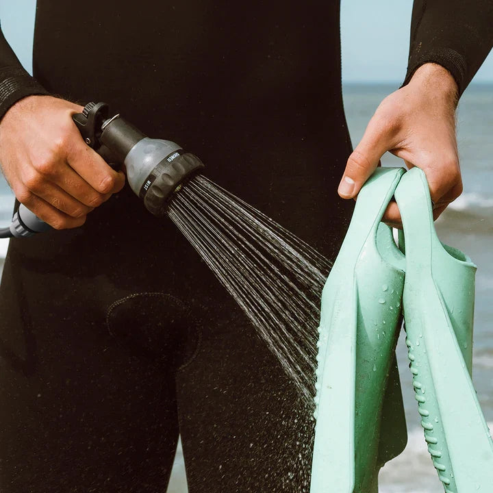 Surfer in a wetsuit rinsing green swim fins with a BeachBox shower gun by the sea