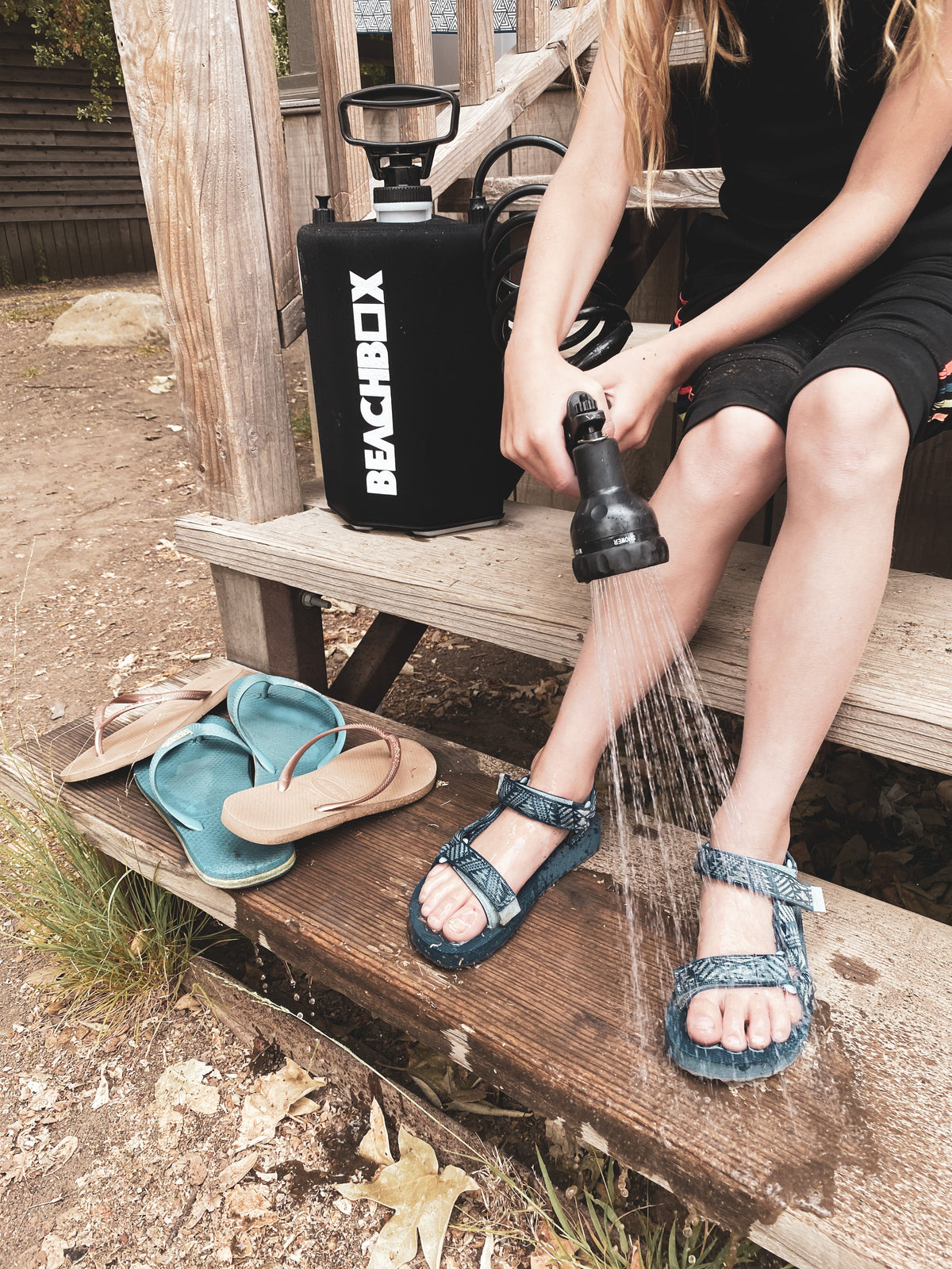 Person rinsing sandy sandals and feet with a BeachBox shower tank on wooden steps