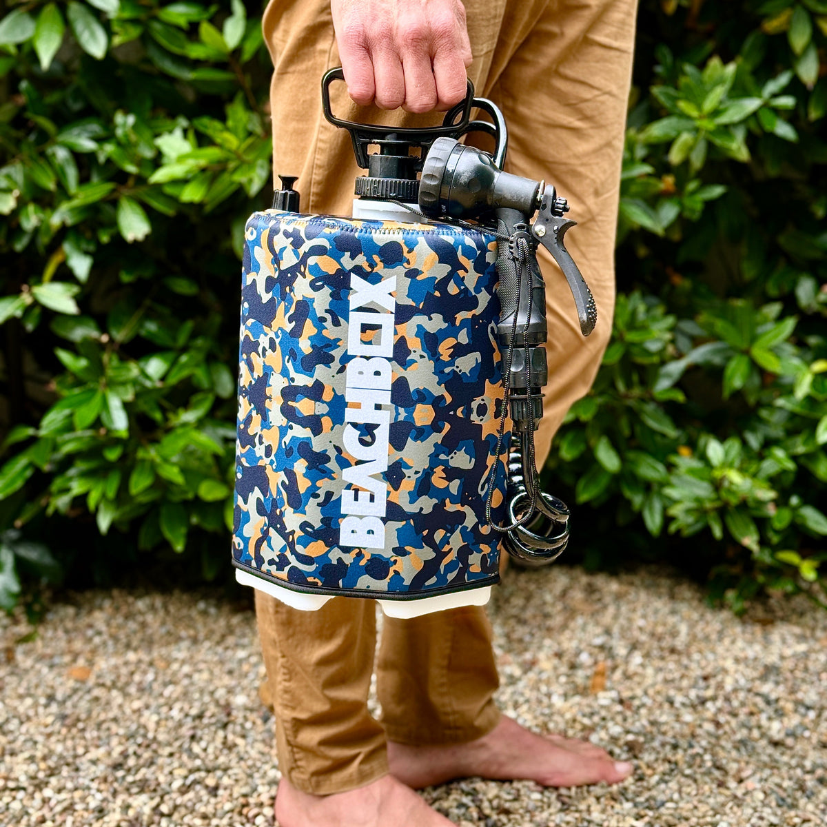 Person carrying a camo BeachBox portable shower tank by its handle with the spray gun attached