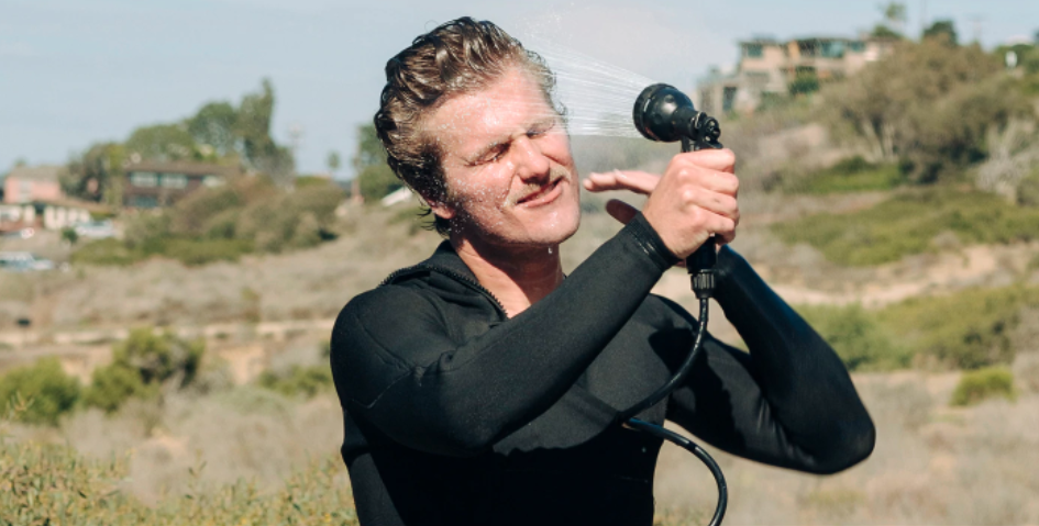 Smiling surfer in a wetsuit rinsing his hair outdoors with a BeachBox shower gun