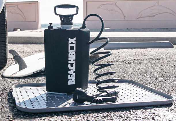Black BeachBox shower tank on a non-slip changing mat beside a surfboard on tarmac
