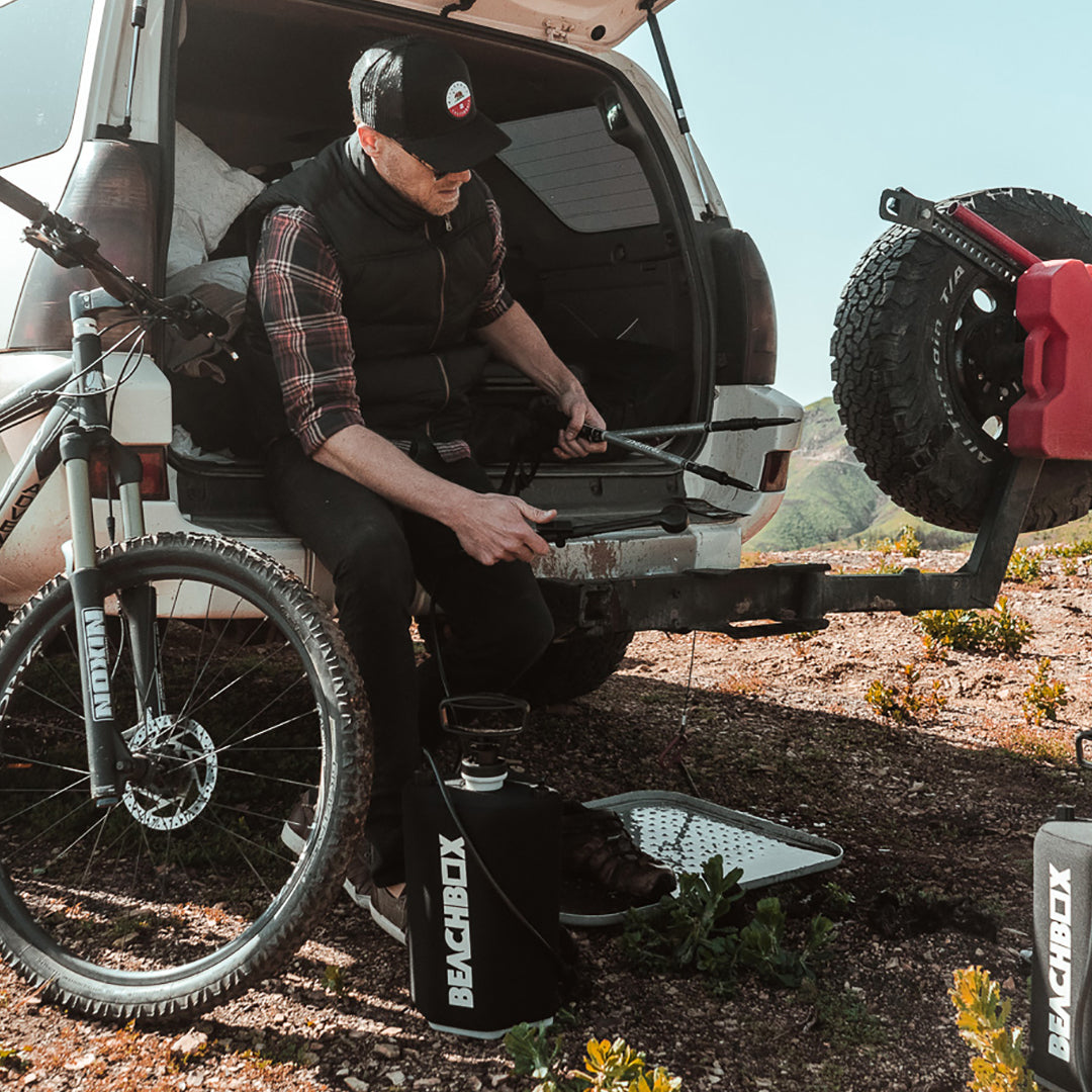 Mountain biker sitting on a truck tailgate with a BeachBox shower tank and mat beside him