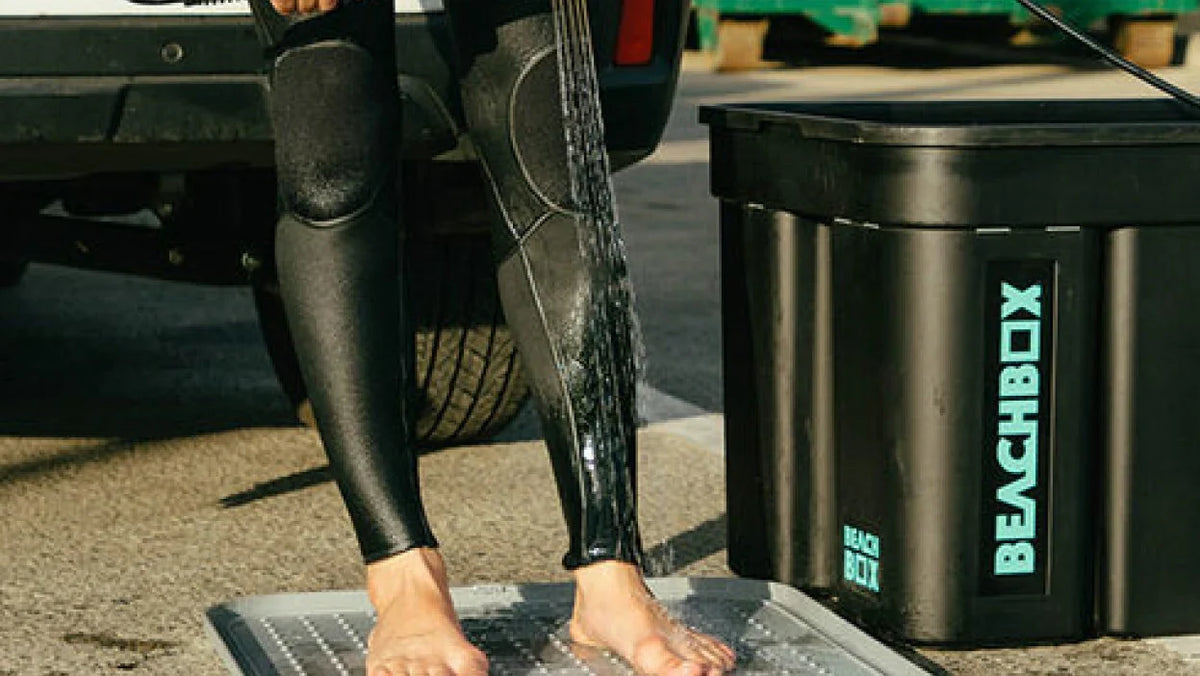Surfer rinsing their legs on a mat beside a BeachBox storage box at a car park