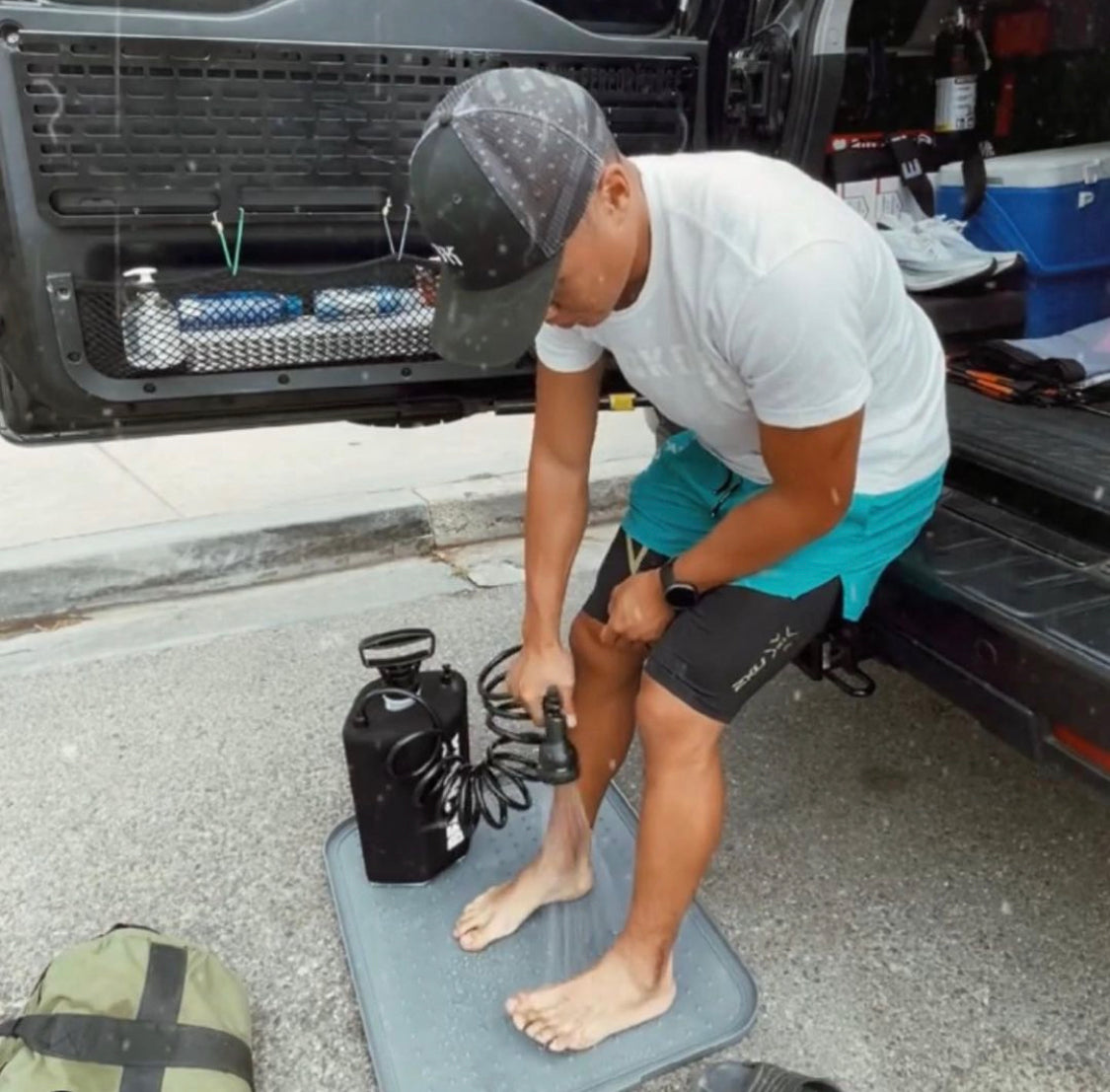 Man rinsing his leg with a BeachBox shower tank on a mat at the back of a truck