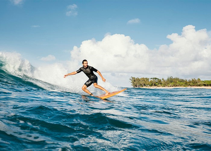 Surfer in a black wetsuit riding a small wave on a longboard near a green headland